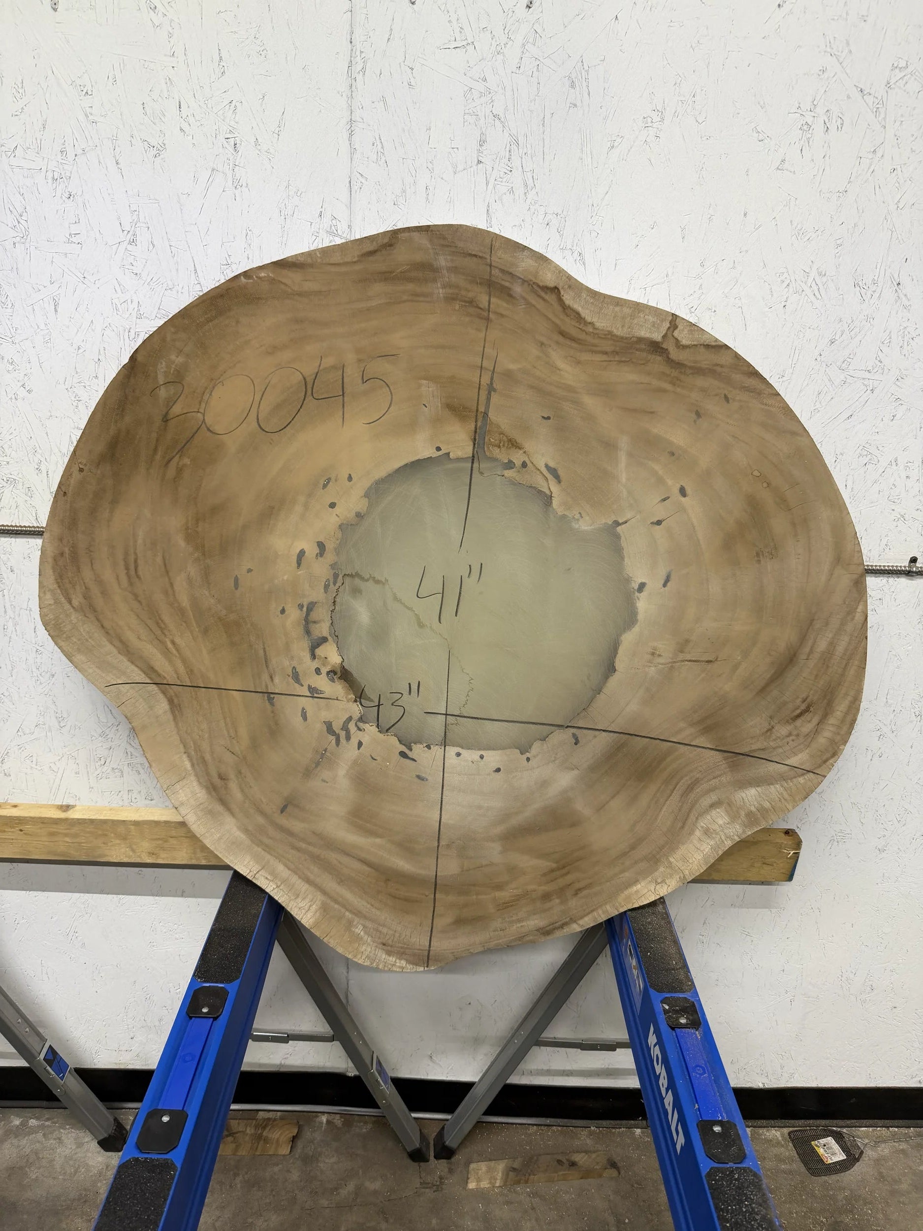 Round Parota live edge wood slab with a central cutout, leaning against a white brick wall on a wooden floor.

41x43