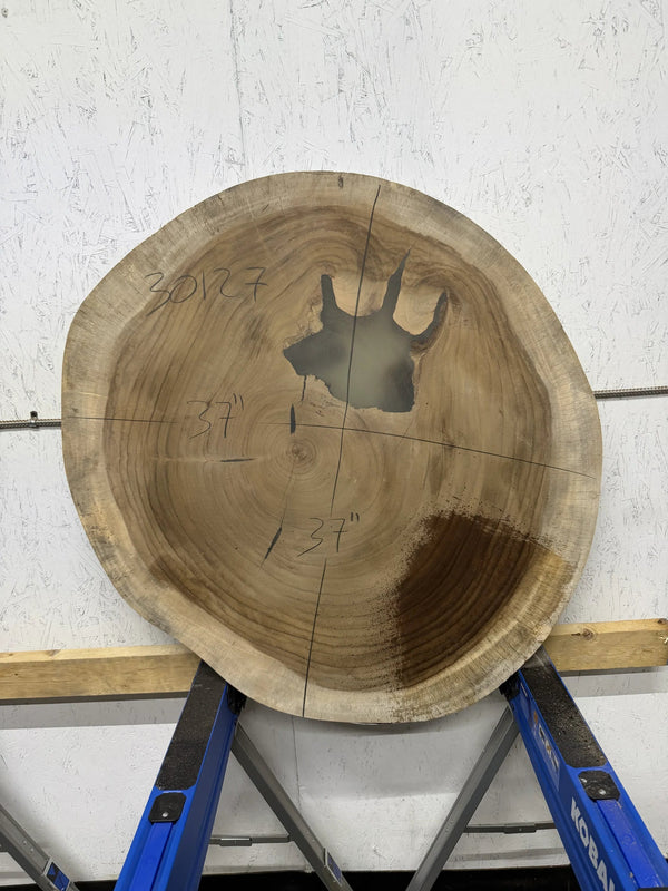 Round Parota live edge wood slab with a central cutout, leaning against a white brick wall on a wooden floor.

